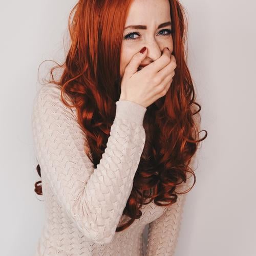 Frau mit langen roten lockigen Haaren lacht in Kamera schauend und hält sich dabei eine Hand vor den Mund Haare & Frisuren lange Haare rote Haare lockiges haar
