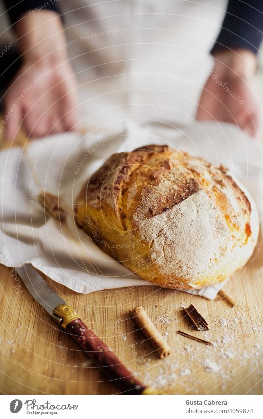Handwerkliches Brot auf einem Holzbrett mit einem Messer, Zimtstückchen und ein wenig Mehl, mit einer unscharfen Person im Hintergrund. Bäcker Bäckerei