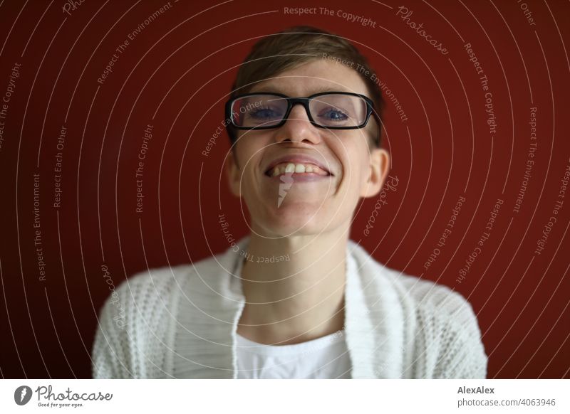 Nahes Portrait einer lächelnden jungen Frau vor roter Wand junge Frau schlank Jeans Top Brille sitzt Schultern Haut groß sportlich gelenkig drinnen Raum Studio