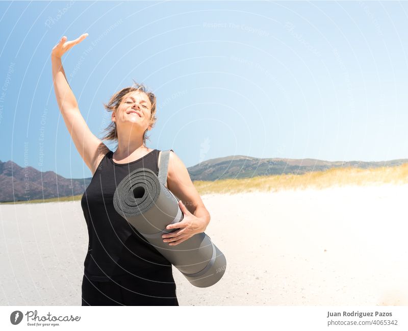 Frau in Sportkleidung genießt am Strand mit einer zusammengerollten Matte unter dem Arm Energie Fitness Pilates aktiv trainiert. Training Unterlage sonnig