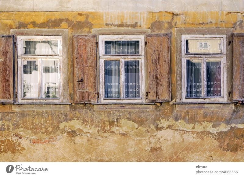 In den drei alten Holzfenstern mit den geöffneten Fensterläden, den blickverdeckenden Gardinen und dem ringsherum abblätternden Putz spiegelt sich die gegenüberliegende Straßenseite