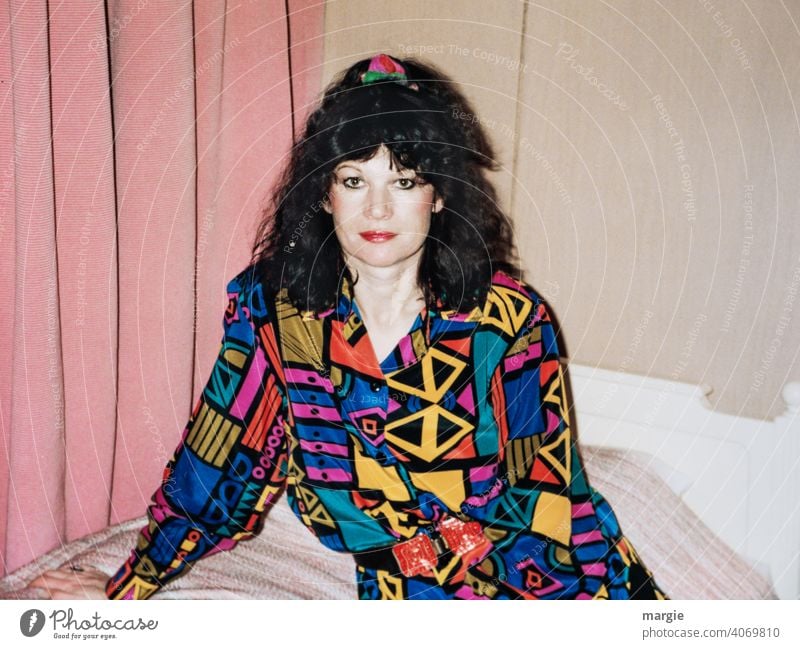 Eine junge Frau sitzt auf einem Bett und schaut in die Kamera Junge Frau bunt Bluse dunkelhaarig Vorhang Fotografie analog analoge fotografie Papier Locken