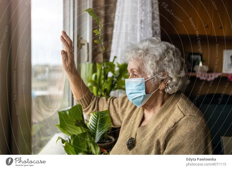 Ältere Frau mit Gesichtsmaske schaut aus dem Fenster zu Hause COVID Sperrung Coronavirus Blick Quarantäne Blick durch das Fenster soziale Distanzierung Glas