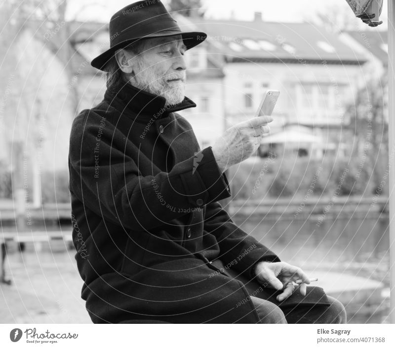 Älterer Herr schaut gebannt auf sein Smartphones Mann Ein Mann allein Außenaufnahme Wasseroberfläche Schwarzweißfoto Schwache Tiefenschärfe Senior sitzend