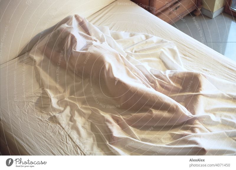 Junge Frau liegt unter einer halb durchsichtigen Decke nackt auf einem Bett junge Frau schlank sportlich gelenkig drinnen Raum hell Tageslicht ästhetisch