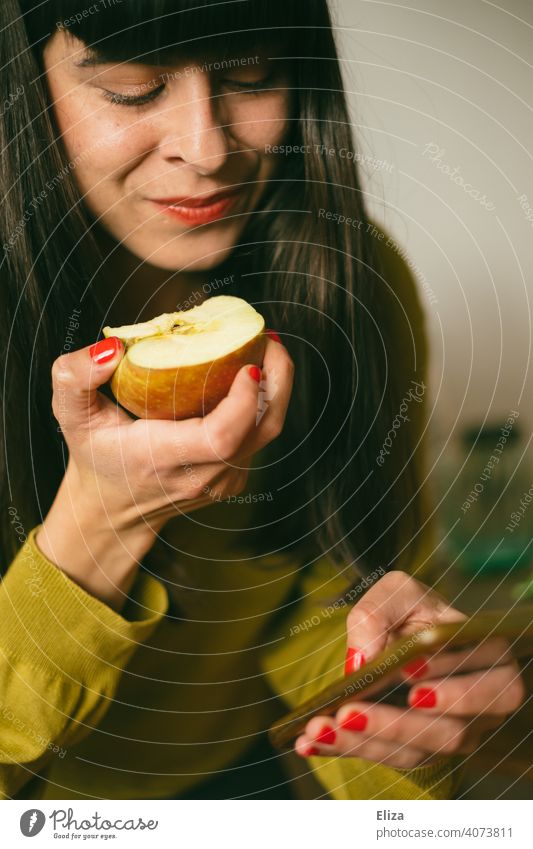 Eine Frau isst einen Apfel, während sie mit ihrem Smartphone beschäftigt ist Telefon Handy Essen gesunde Ernährung Obst Snack snacken online nebenbei