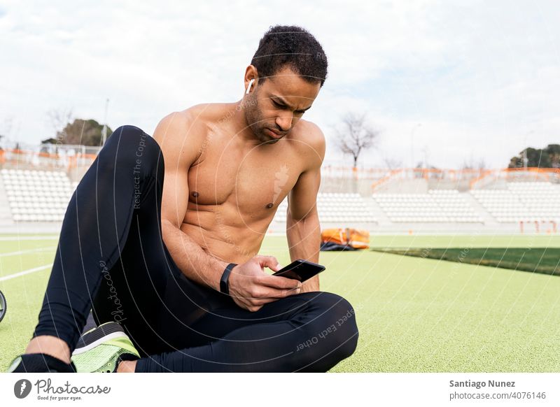 Vorderansicht eines Athleten, der das Telefon in der Leichtathletikbahn benutzt Sportbahn per Telefon Sitzen Schwarzer Mann ruhen aussruhen Stock Hintergrund