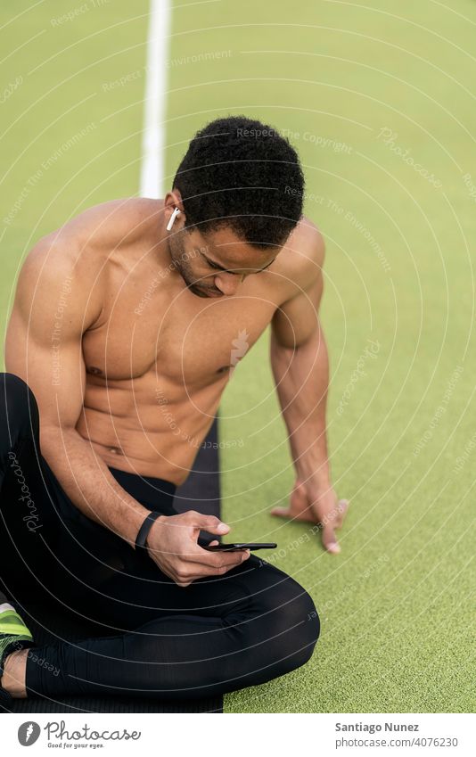 Seitenansicht eines Athleten, der das Telefon in der Leichtathletikbahn benutzt Sportbahn per Telefon Sitzen Schwarzer Mann ruhen aussruhen Stock Hintergrund