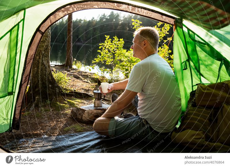 Mann macht Kaffee mit Espressomaschine auf Lagerfeuer im Wald am Ufer eines Sees, sitzt im Zelt, macht ein Feuer, Grillen. Glückliche Isolation Konzept. Finnland erforschen. Skandinavische Landschaft.