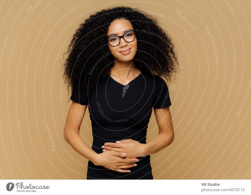 Erfreute afroamerikanische Frau mit lockigem Haar, hält die Hände auf dem Bauch, fühlt sich gut, nachdem sie nahrhaftes leckeres Abendessen gegessen hat, schaut glücklich in die Kamera. Junge erfreute Frau erfährt von der Schwangerschaft.