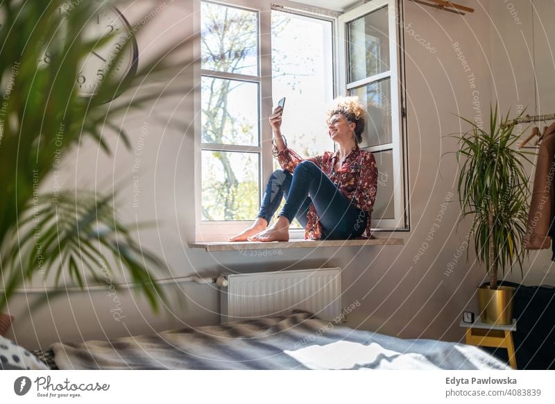 Junge Frau sitzt auf dem Fensterbrett und verwendet Smartphone Glück Lächeln heiter Appartement Freizeit Schlafzimmer Haus heimwärts Raum allein eine Menschen