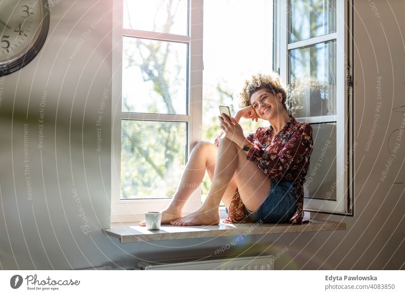 Junge Frau sitzt auf dem Fensterbrett und verwendet Smartphone Glück Lächeln heiter Appartement Freizeit Schlafzimmer Haus heimwärts Raum allein eine Menschen