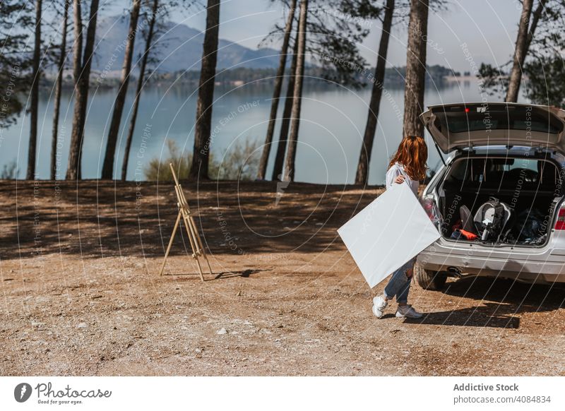 Anonyme Künstlerin mit Leinwand in der Nähe von Auto Landschaft PKW See Kofferraum offen Staffelei Frau blanko modern Hobby Lifestyle Freizeit Natur sonnig