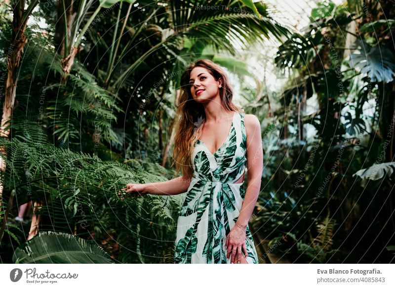 Porträt der schönen blonden jungen Frau lächelnd bei Sonnenuntergang in einem grünen Haus, umgeben von tropischen Pflanzen. Glück und Lifestyle-Konzept