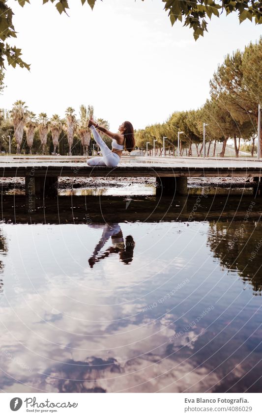 junge schöne asiatische Frau macht Yoga in einem Park. Sitzen auf der Brücke mit Reflexion auf dem Wasser See. Yoga und gesunder Lebensstil Konzept praktizieren
