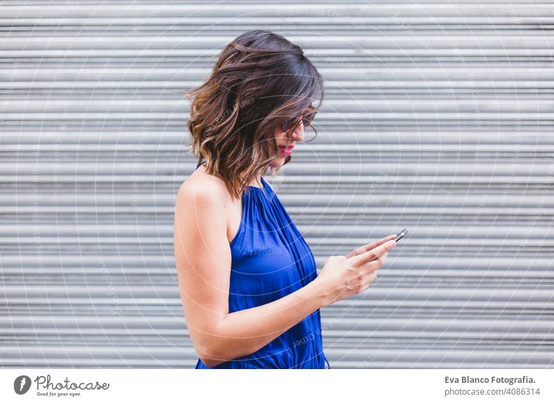 junge schöne Frau mit Handy und lächelnd. Sie ist auf der Straße, trägt ein lässiges blaues Kleid, Sonnenbrille und lächelnd. LIfestyle im Freien, modernes Leben und Technologie