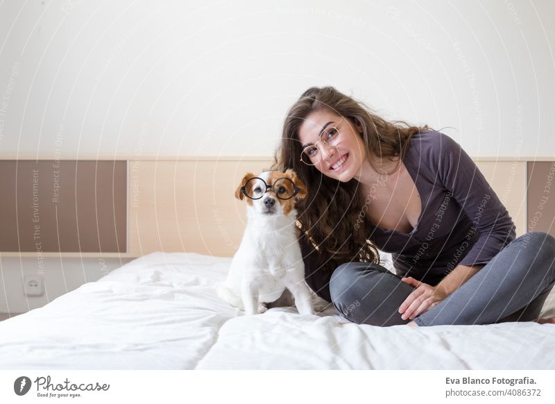 schöne junge Frau auf dem Bett liegend mit ihrem niedlichen kleinen Hund daneben. Zuhause, drinnen und Lifestyle. Sie tragen eine Hipster-Brille gemütlich müde