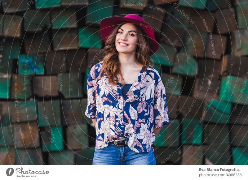 Porträt einer jungen schönen Frau, die lässige Kleidung und einen modernen Hut trägt, über grünen Holzblöcken Hintergrund steht und lächelt. Lebensstil im Freien.