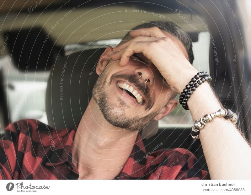 junger mann sitzt lachend im auto portrait 30 jahre hand armreif zaehne fahrersitz happy positiv sonne mode urlaub holilday relaxed entspannt symphatisch