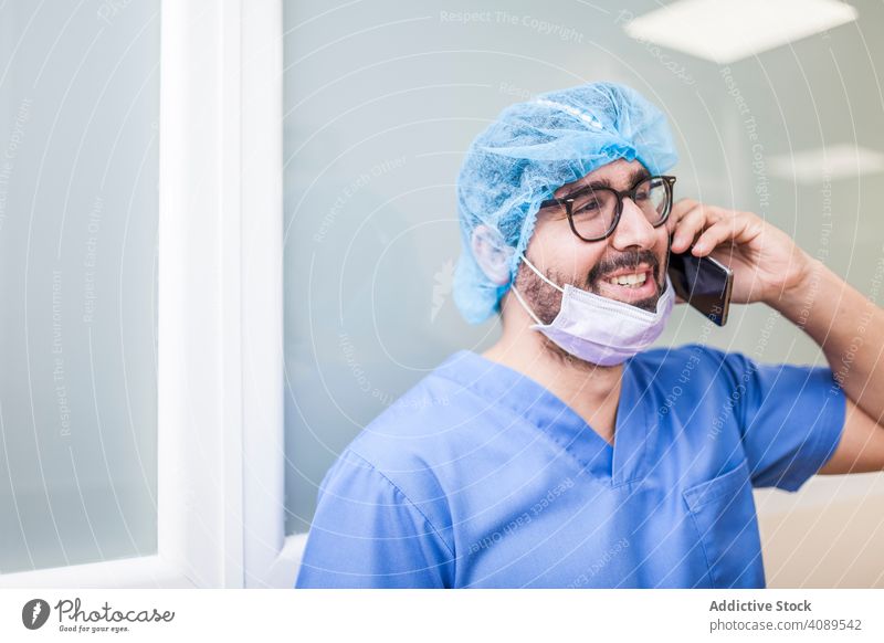 männliches OP-Personal, das sein Smartphone benutzt Job Körperhaltung überdrüssig im Innenbereich Hut Wand Krankenhaus Operationssaal professionell Flur weiß