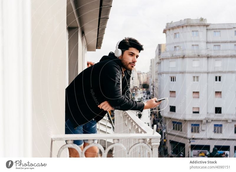 Junger glücklicher Mann hält Smartphone auf Balkon Glück Kopfhörer Headset Lehnen Reling Terrasse jung männlich Lächeln Freude heiter gutaussehend gelungen cool