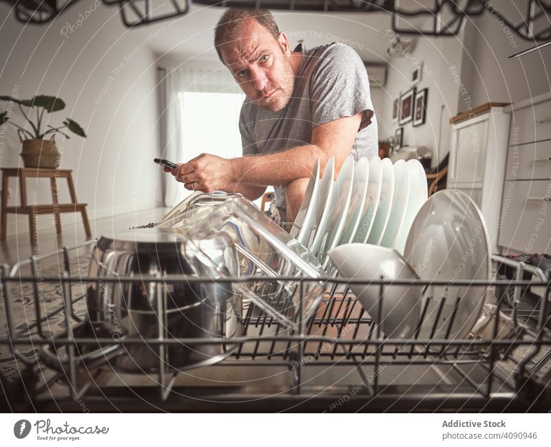 Wütender erwachsener Mann mit Smartphone beim Reparieren der Spülmaschine in der Küche Vater fixieren Geschirrspüler Werkzeuge wütend Reparatur Familie