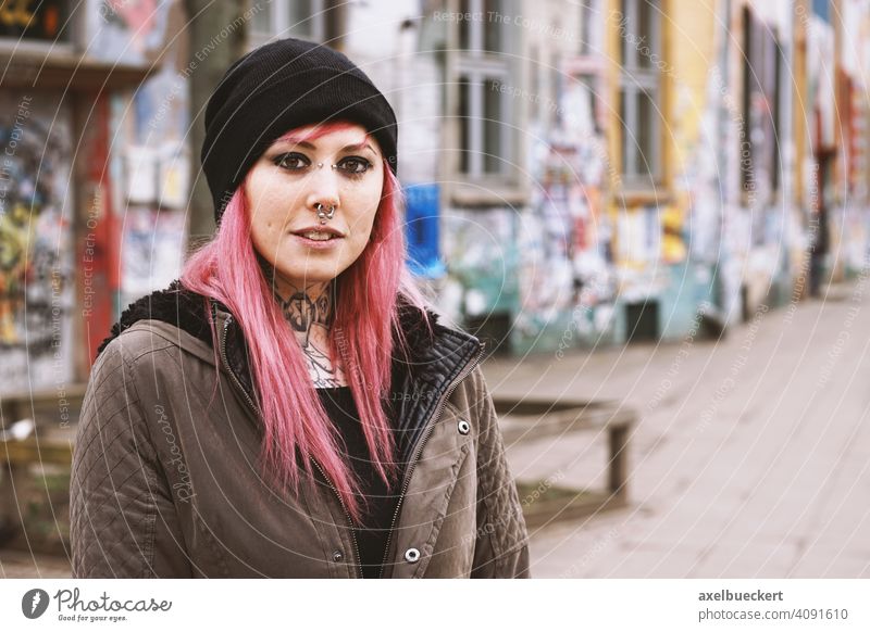 junge Frau mit pinken Haaren, Piercings und Tattoos vor Graffiti beschmierten Häusern Junge Frau echte Menschen Subkultur Haus pinke haare gepierct tätowiert