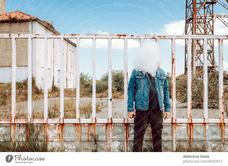 Anonymer Vaper stehend in der Nähe von rostigen Barriere Mann Raps Cloud Zaun verfallen Gebäude industriell ausatmen Habitus männlich Hipster E-Zigarette Dampf