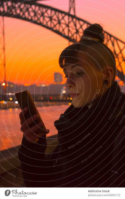 Stilvolle Frau benutzt Smartphone am Abend am Ufer Sonnenuntergang benutzend Stauanlage Großstadt Fluss stylisch jung hell alternativ Generation Subkultur
