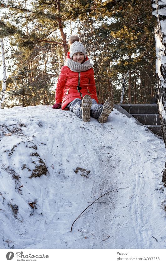 Junges lächelndes süßes Mädchen in einem hellen Sportanzug bereitet sich darauf vor, den Hügel hinunter zu schlittern. Glückliche Kindheit, Winter Outdoor-Aktivität. Vertikale Aufnahme