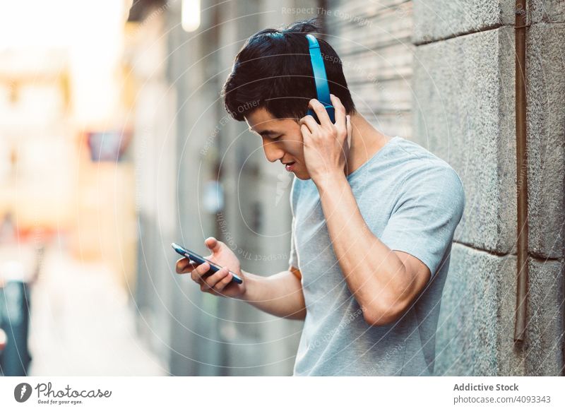 Inhalt Asiatischer Mann mit Kopfhörern hört auf der Straße Musik auf seinem Smartphone Großstadt zuhören benutzend Streaming cool freudig modern lässig stehen