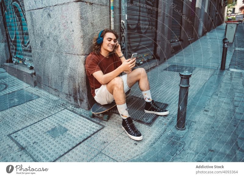 Entspannter Mann mit Kopfhörern, der sein Smartphone benutzt, während er sich auf einem Longboard auf der Straße ausruht benutzend entspannt sorgenfrei