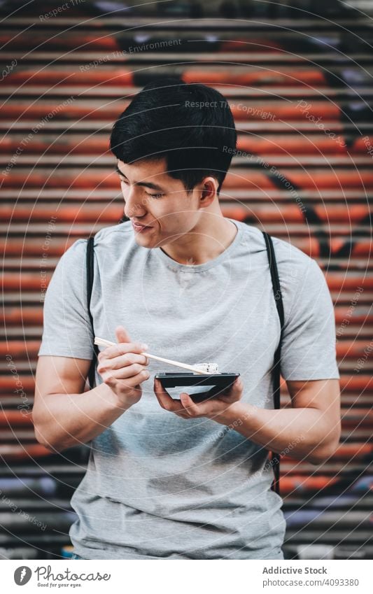 Asiatischer Mann isst Essen zum Mitnehmen mit Stäbchen auf einer Straße in der Stadt Imbissbude Essstäbchen Straßenessen hungrig Großstadt Asiatische Küche