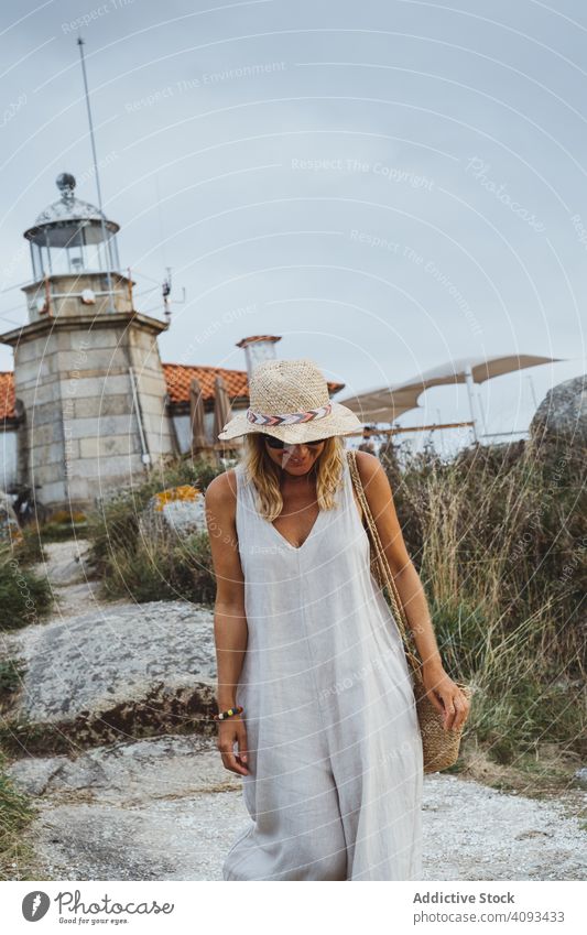 Frau mit Hut steht vor einem Leuchtturm am Meeresufer Leuchtfeuer Stein Küste Felsen Architektur Spaziergang Gebäude Gras reisen Ausflug trist Urlaub Cloud