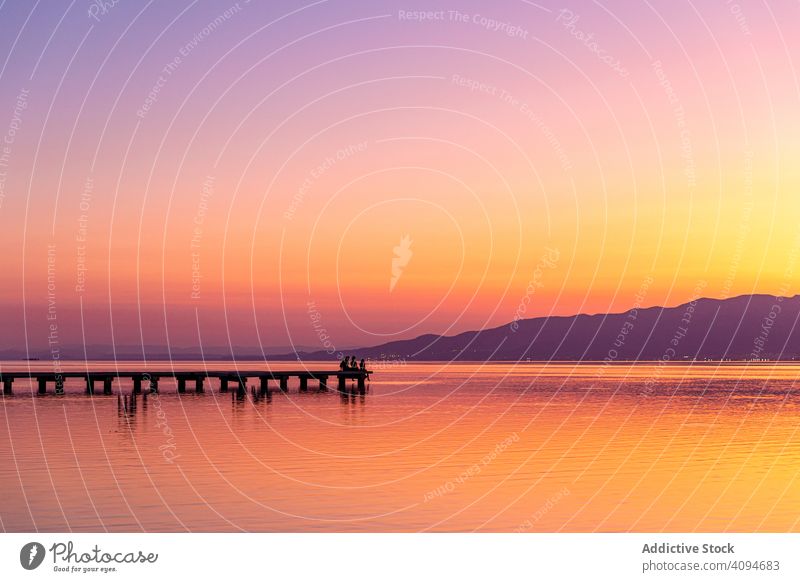 Anonyme Touristen, die bei Sonnenuntergang auf dem Steg sitzen Pier See Reisender Silhouette ruhig wolkenlos Himmel Abend hell Wasser Tourismus Ausflug