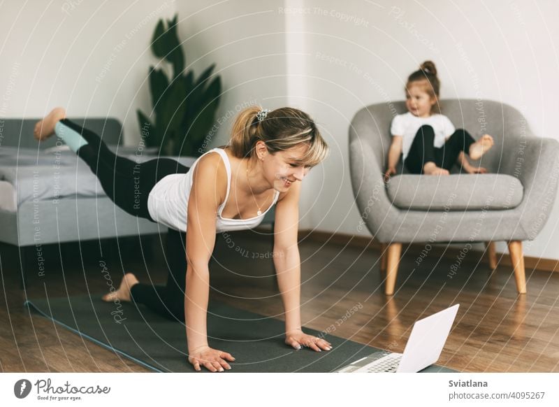 Eine junge Mutter treibt zu Hause Sport und schaut sich eine Video-Fitnessstunde auf einem Laptop an, und ihr kleines Baby sitzt auf einem Stuhl. Heimtraining, Fitness, Sport