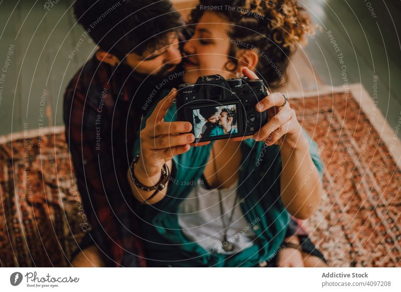 Ein fröhliches Paar umarmt sich und nimmt sich zu Hause vor der Kamera Küssen Fotokamera Selfie Hipster freudig Kuss Zuneigung umarmend benutzend sitzen ruhen