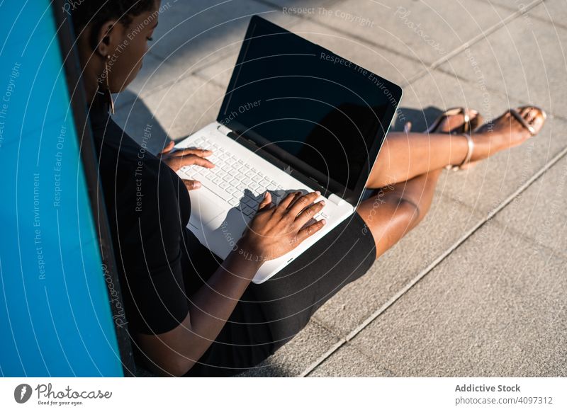 Seriöse Frau surft am Laptop in der Nähe eines modernen Gebäudes sich[Akk] entspannen konzentriert ernst benutzend Surfen Afroamerikaner elegant Straße Kleid