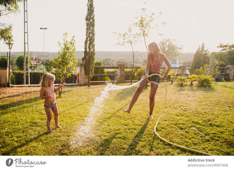 Teenager-Mädchen besprüht kleine Schwester im Garten Spaß Wasser Rasen Spray Schlauch sonnig tagsüber Hof Zusammensein Kind Glück heiter spielen Urlaub