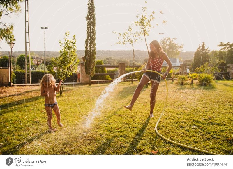 Teenager-Mädchen besprüht kleine Schwester im Garten Spaß Wasser Rasen Spray Schlauch sonnig tagsüber Hof Zusammensein Kind Glück heiter spielen Urlaub