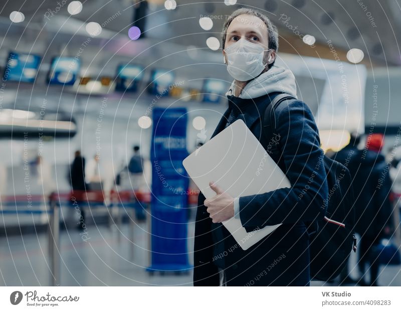 Männlicher Reisender trägt medizinische Einweg-Schutzmaske im Flughafen, kehrt aus dem Ausland zurück, wo sich das Coronavirus ausbreitet, trägt Rucksack, kümmert sich um Gesundheit, schützt sich vor Virus, hat Flug verschoben
