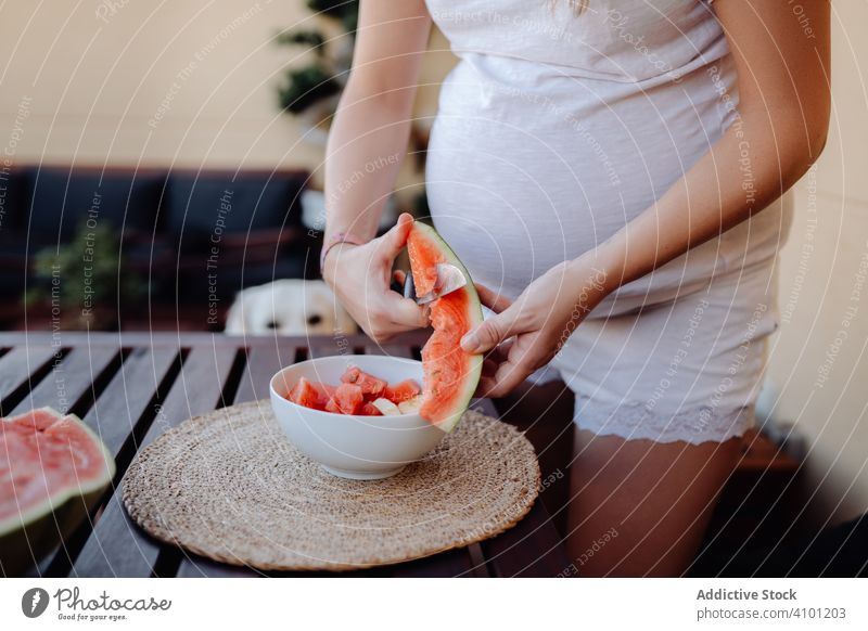 Gesichtslose schwangere Frau, die eine Wassermelone in eine Schüssel schneidet Sommer geschnitten Erwartung Tisch Terrasse zuschauen Mutterschaft