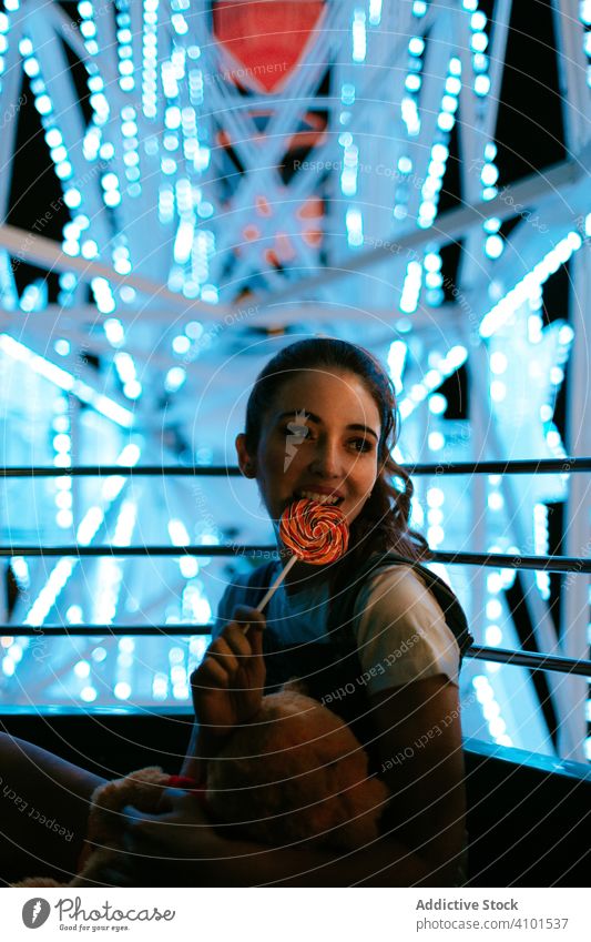 Millennial-Frau sitzt im Riesenrad und isst Lollipop Karussell Vergnügungspark süß heiter Nacht Teenager tausendjährig Bonbon Spielzeug Glück positiv freudig