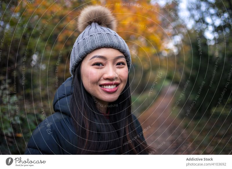 Lächelnde asiatische Frau in warmer Kleidung in einem Park mit Bäumen und steinigem Zaun stehend Garten stylisch Herbst Natur Straße Baum Weg Blätter Hut