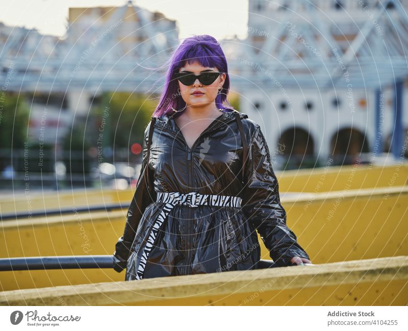 Frau mit violetten Haaren in der Nähe von Metallkonstruktionen stylisch urban purpur Frisur Jacke Sonnenbrille glänzend Zaun gelb Mode jung Stil Model Straße