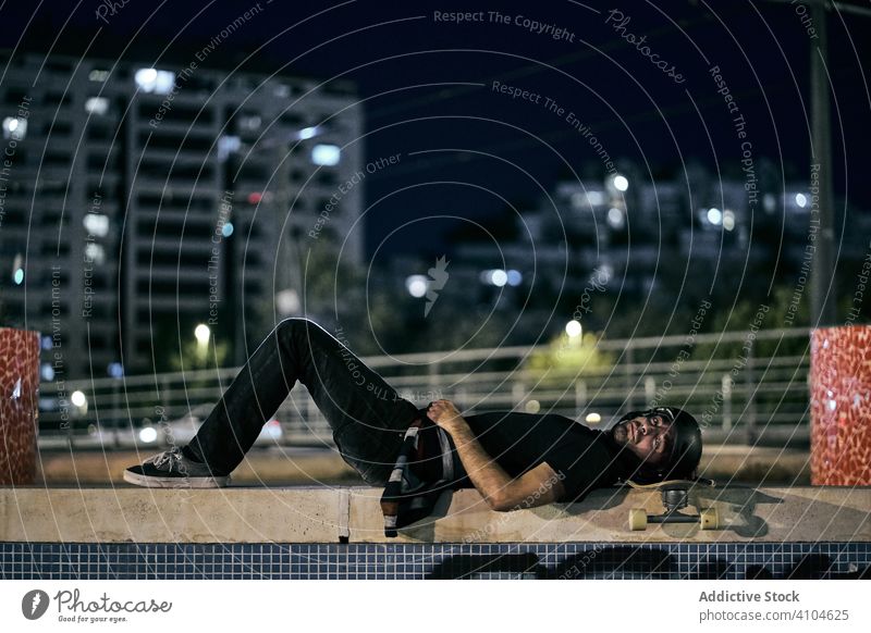 Männlicher Skateboarder liegt nachts auf einem Zaun in der Stadt Mann Großstadt Lügen Nacht ruhen sich[Akk] entspannen Schutzhelm Schlittschuh Straße Abend jung