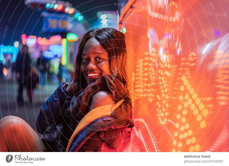 Fröhliche schwarze Frau auf Rummelplatz Jahrmarkt Lächeln Nacht Arkaden Illumination sitzen ruhen Wochenende ethnisch Vergnügen Anziehungskraft farbenfroh