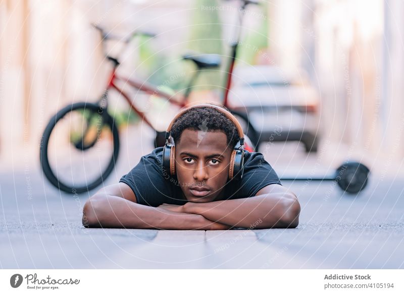 Fokussierter junger schwarzer männlicher Teenager, der auf der Straße Musik hört zuhören Mann Kopfhörer cool Lügen Asphalt Lifestyle Schüler