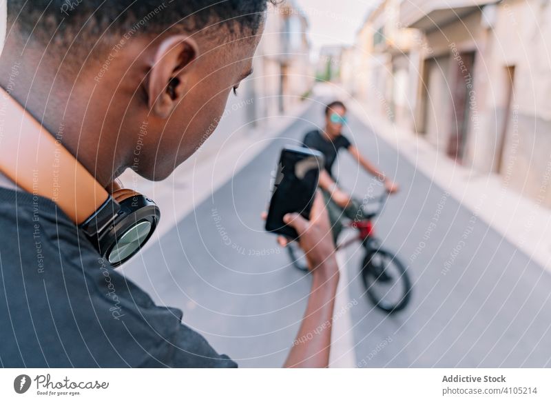 Junger schwarzer männlicher Teenager fotografiert mit seinem Handy einen Freund auf der Straße fotografieren Funktelefon Männer bmx Smartphone cool Fahrrad