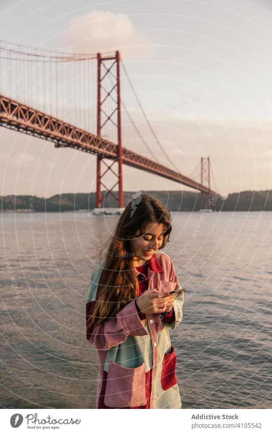 Glückliche Frau surft mit Smartphone gegen Brücke benutzend Handy Nachrichtenübermittlung Freizeit sich[Akk] entspannen Lissabon Portugal Mitteilung Anschluss